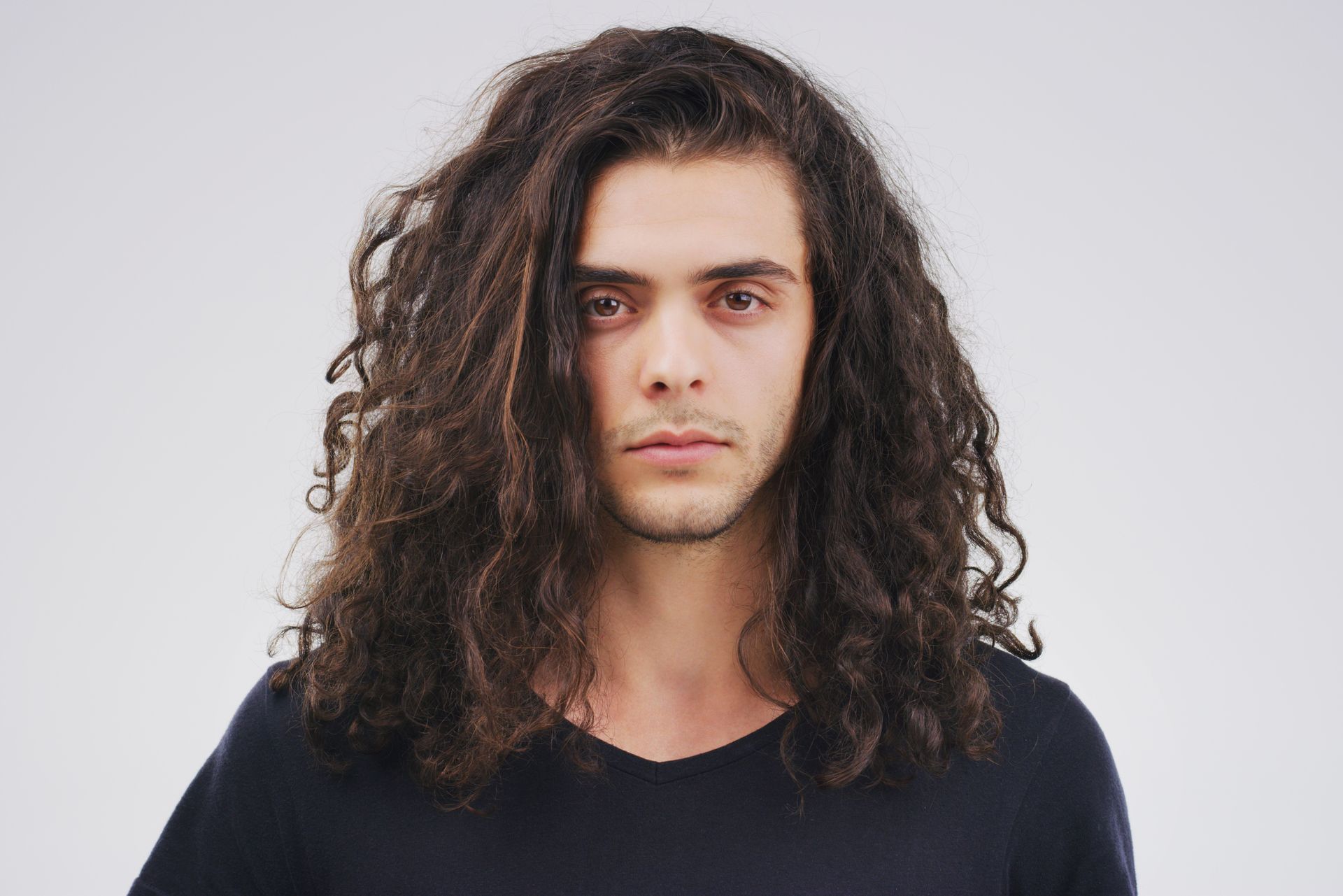 Man with long, curly brown hair stares at the camera, wearing a dark v-neck shirt against a white backdrop.