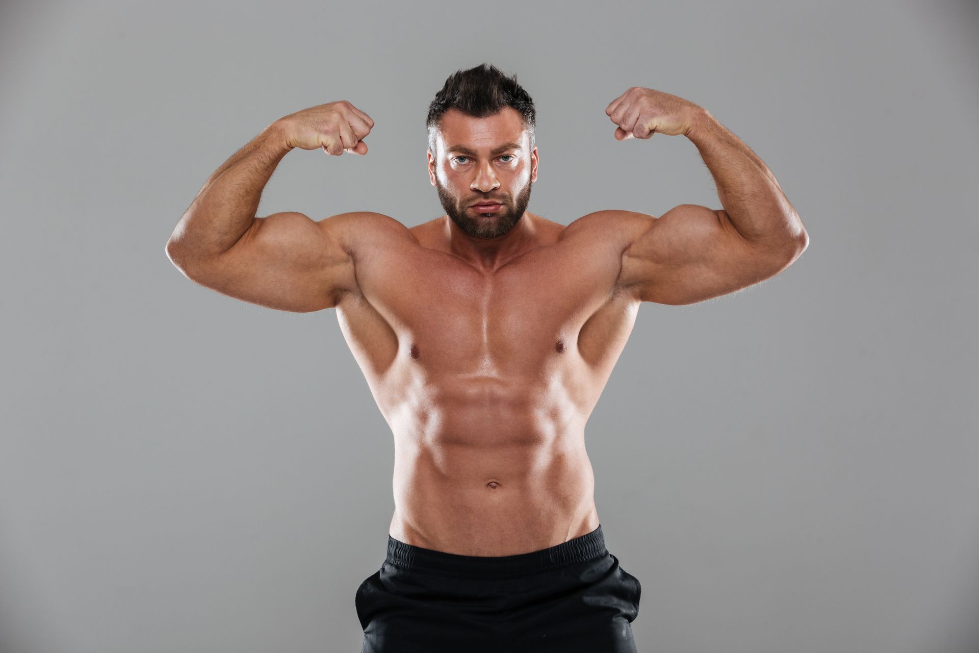 Bodybuilder flexing biceps, shirtless, wearing black shorts, grey background.