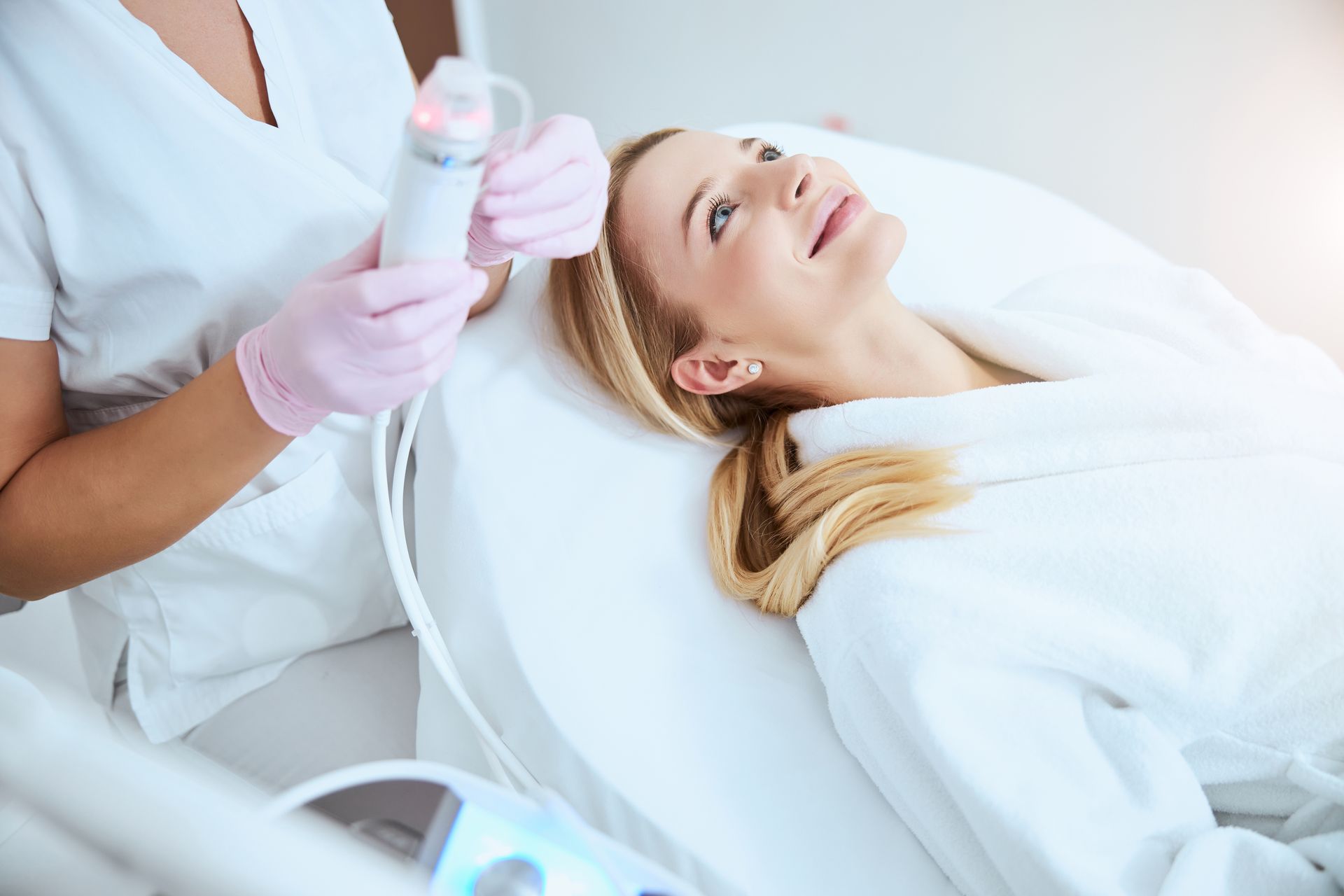 Woman receiving facial treatment in a spa, attended by a gloved aesthetician with a handheld device.