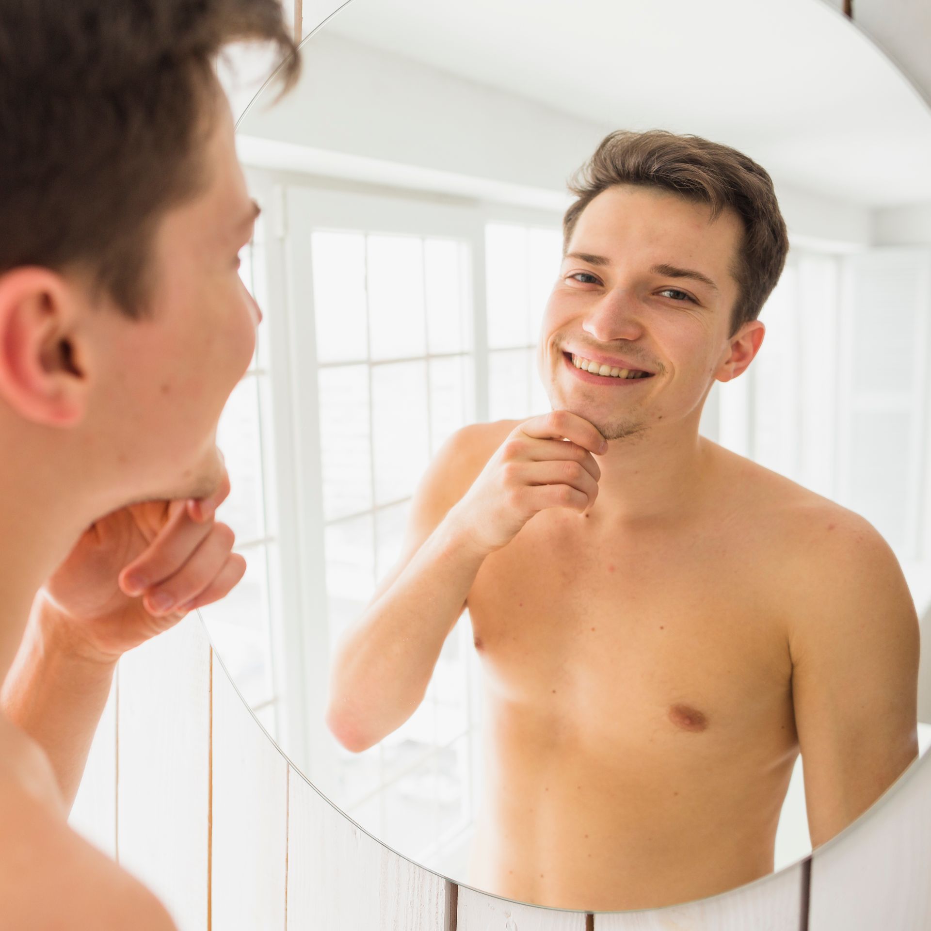 Young man looking in a mirror, smiling and touching his chin. He is shirtless in a bathroom.