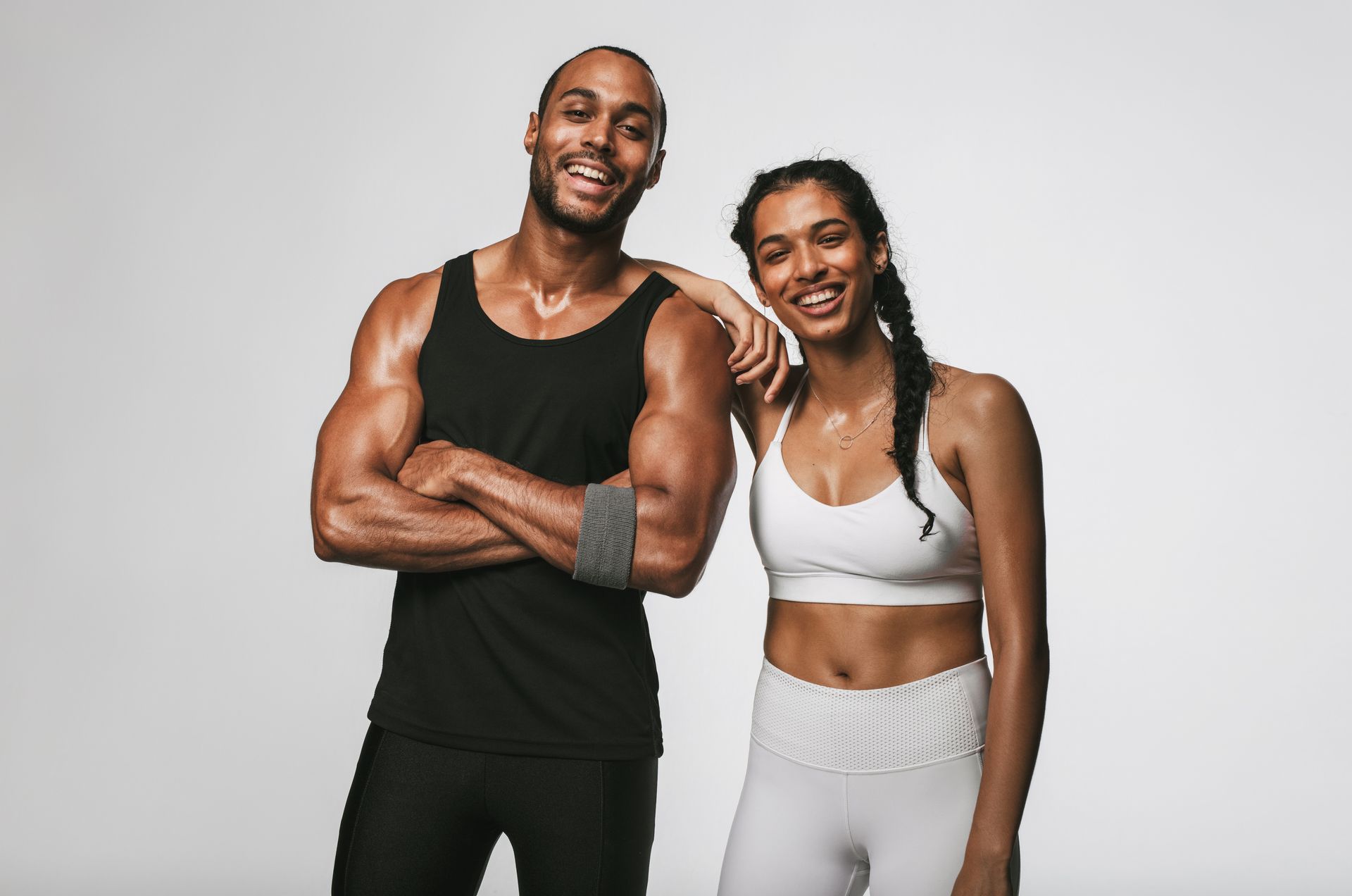 Smiling athletic couple, man in black tank, woman in white sports bra, both with sweat, grey backdrop.