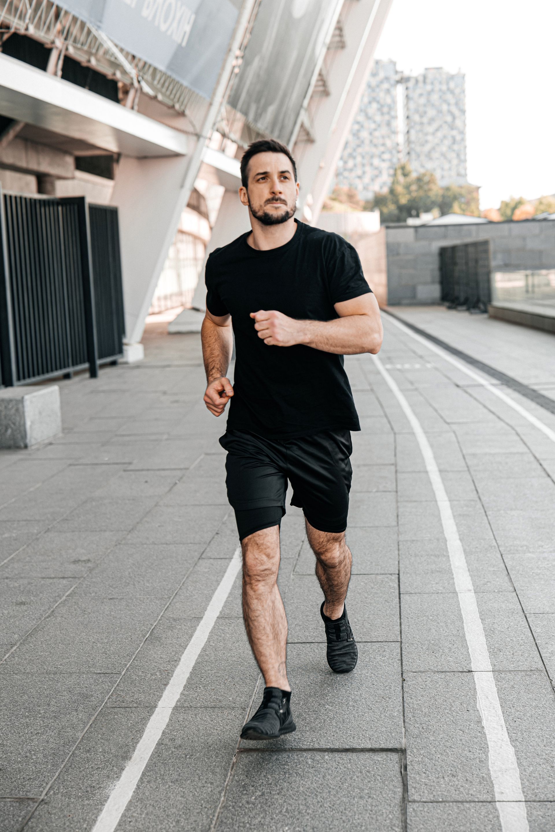 Man in black running shorts and shirt, running on a paved path in an urban setting.