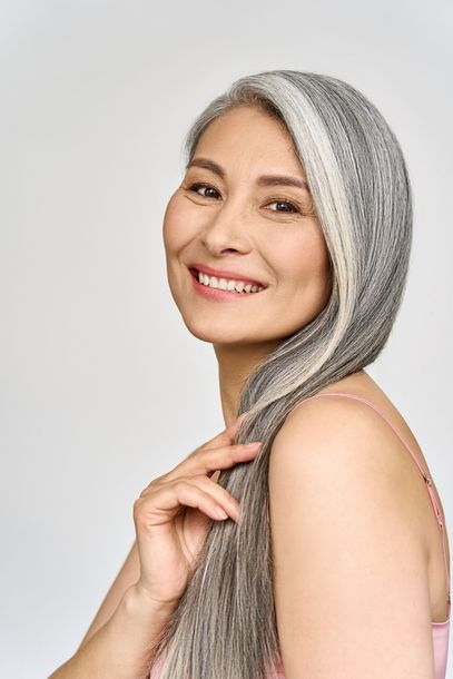 Smiling Asian woman with silver hair, pink top, holding hair, against a white background.