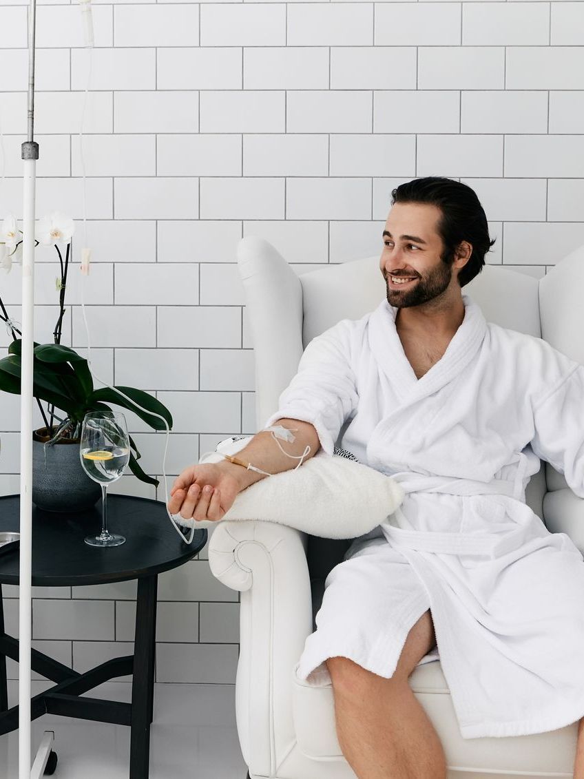 Man in white robe receives IV drip, seated in a white chair, smiling. White tiled background.