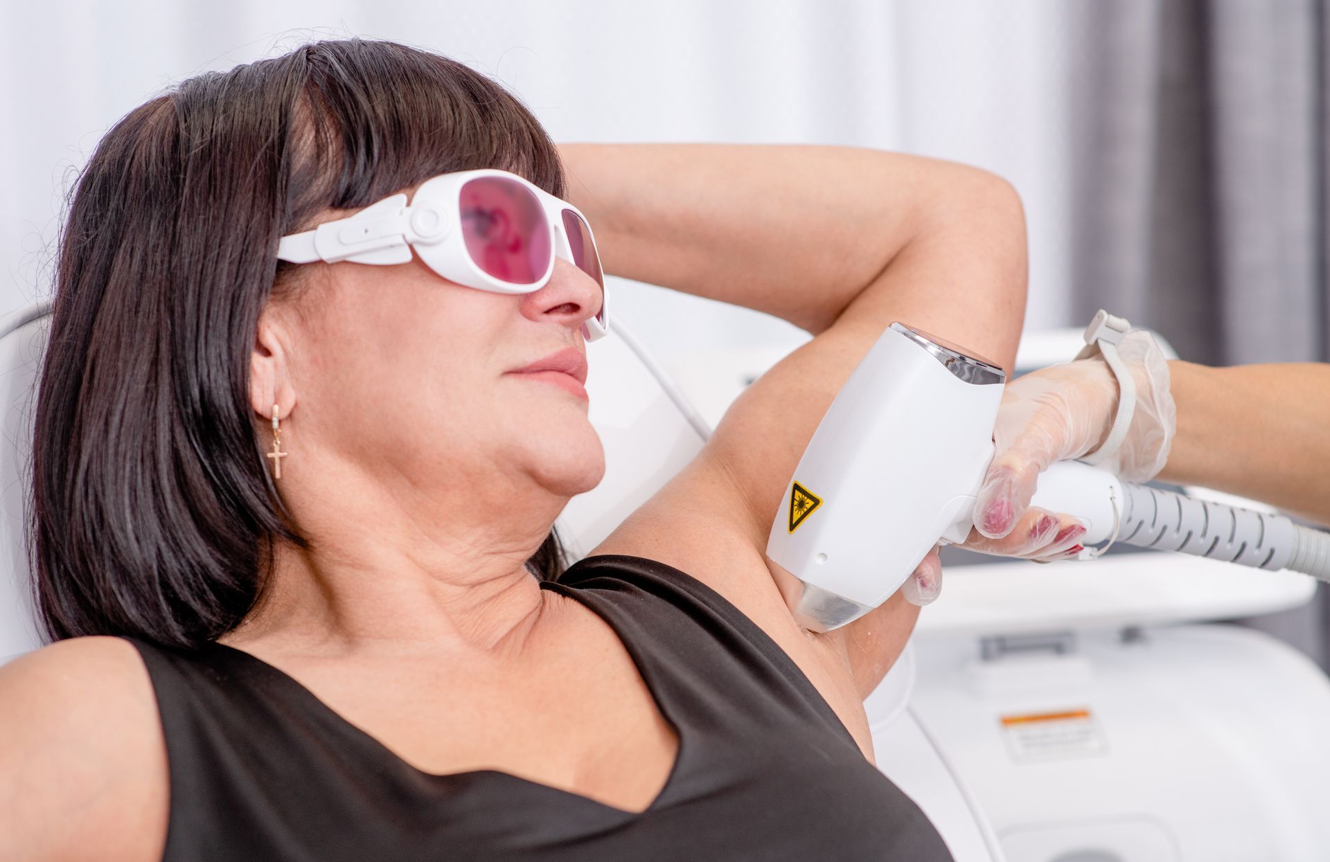 Woman undergoing laser hair removal on her underarm, wearing safety glasses, in a clinic.