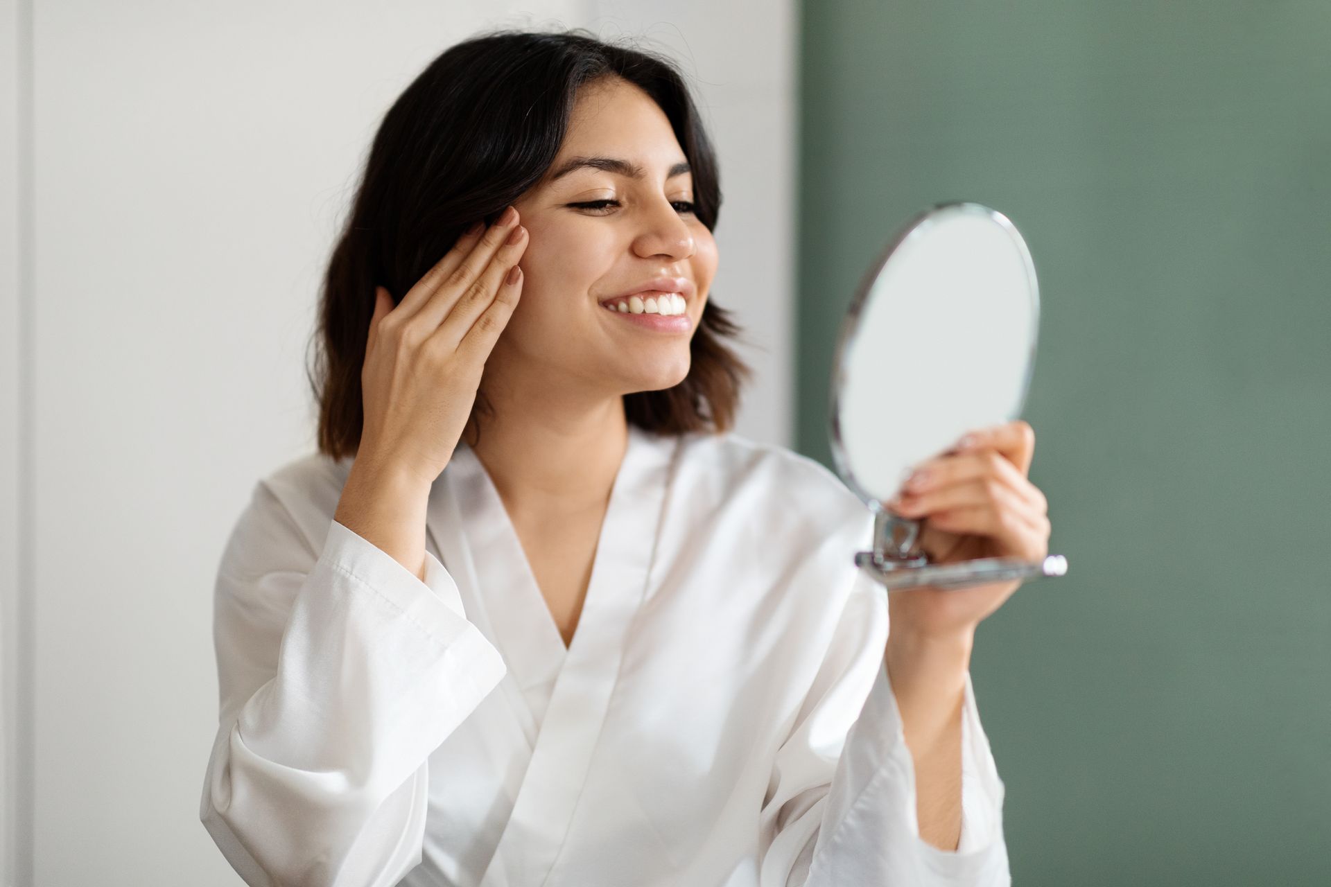 Woman in a white robe smiles while looking at her reflection in a handheld mirror; touching her face in a bathroom.