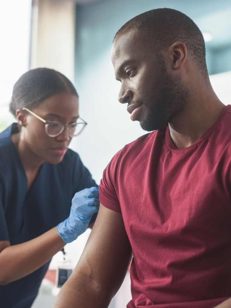 Nurse administering a vaccine to a man's upper arm. Both are Black. The nurse wears blue gloves and scrubs.