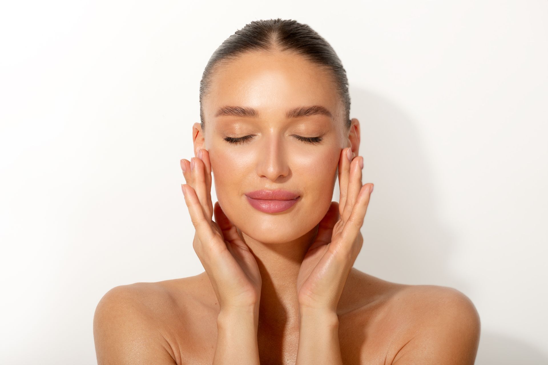 Woman with eyes closed, gently touching her face with hands, white background.