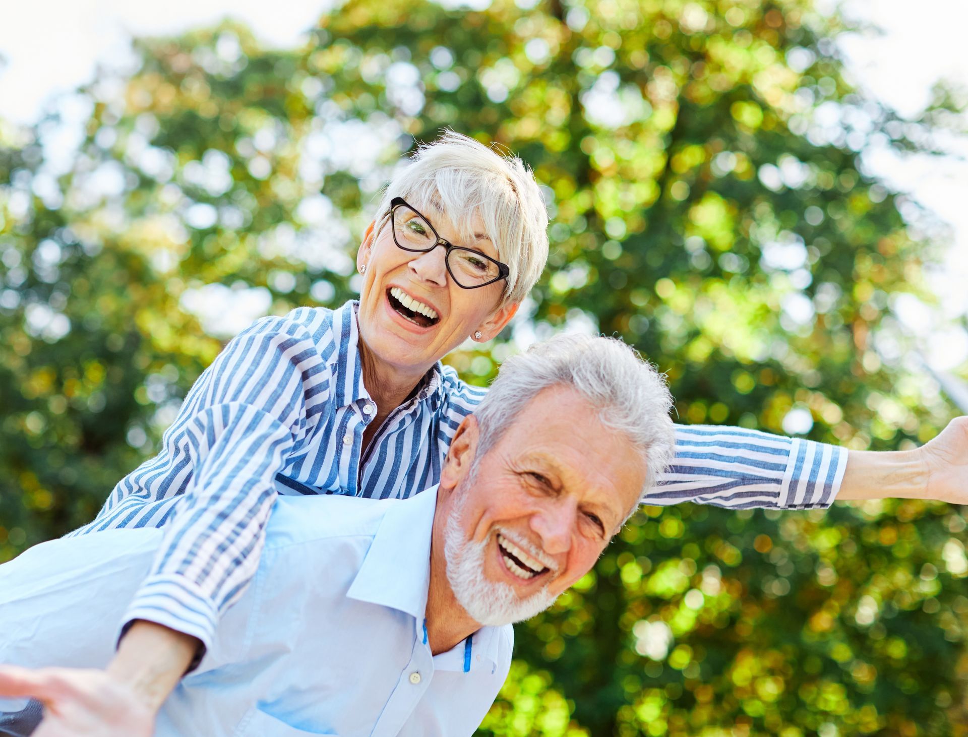 Elderly couple smiling, woman on man's back, outdoors in sunlight.