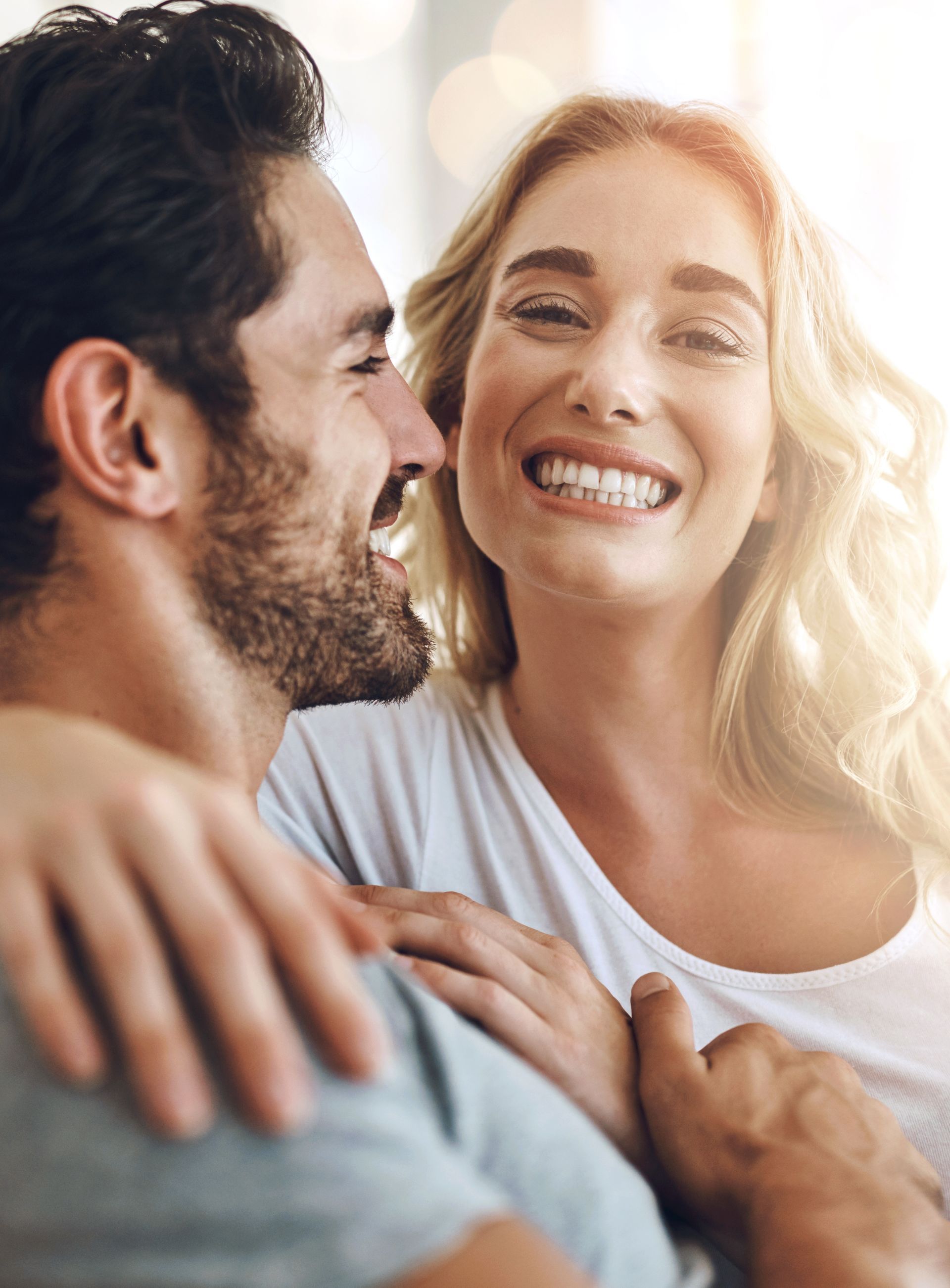 Smiling couple embracing, lit by sunlight. Woman with blonde hair and white shirt, man with dark hair and beard.