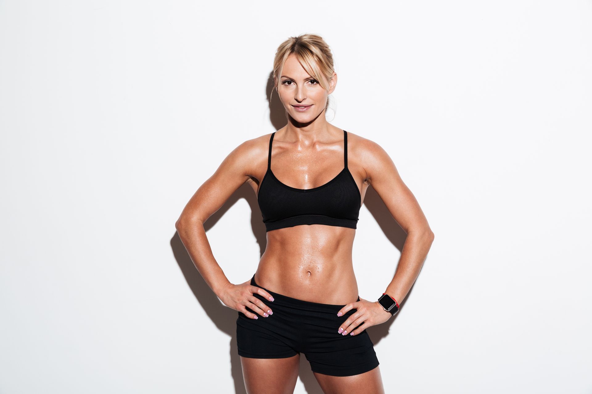 Fit woman in workout clothes, hands on hips, smiling. Sweaty, against a white background.