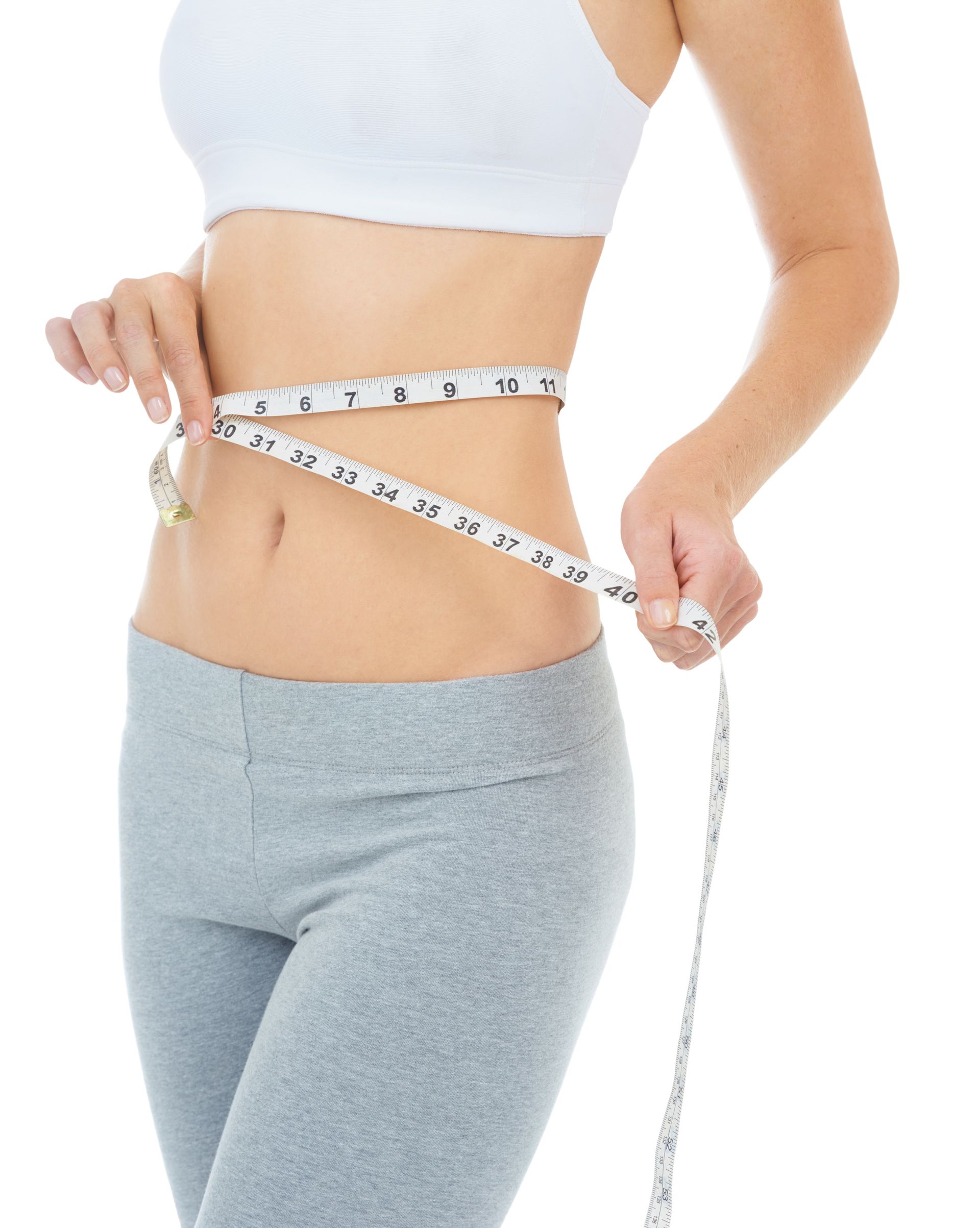 Woman in white sports bra and gray leggings measures her waist with a tape.