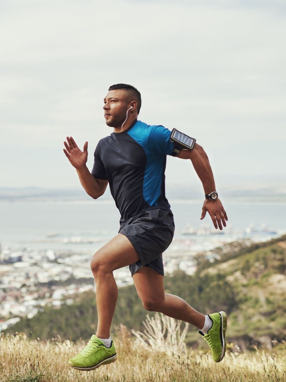 Runner on a hillside, wearing a black and blue shirt, shorts, and green shoes, with a cityscape in the background.