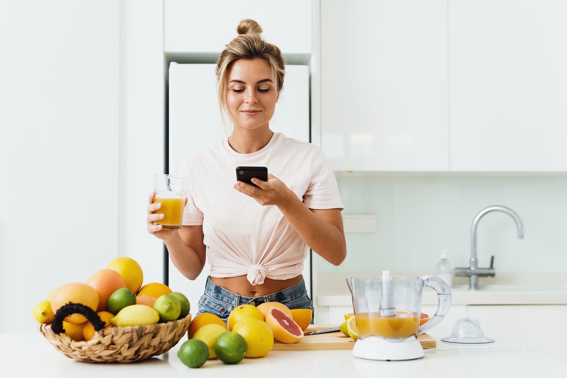 Woman in kitchen, holding juice and phone, surrounded by fruit.