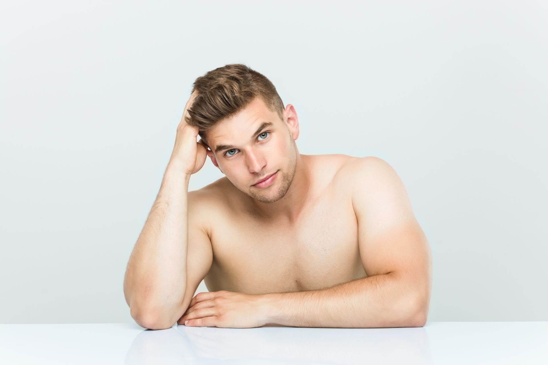 Shirtless man with light skin leans on a table, hand on head, looking thoughtful.