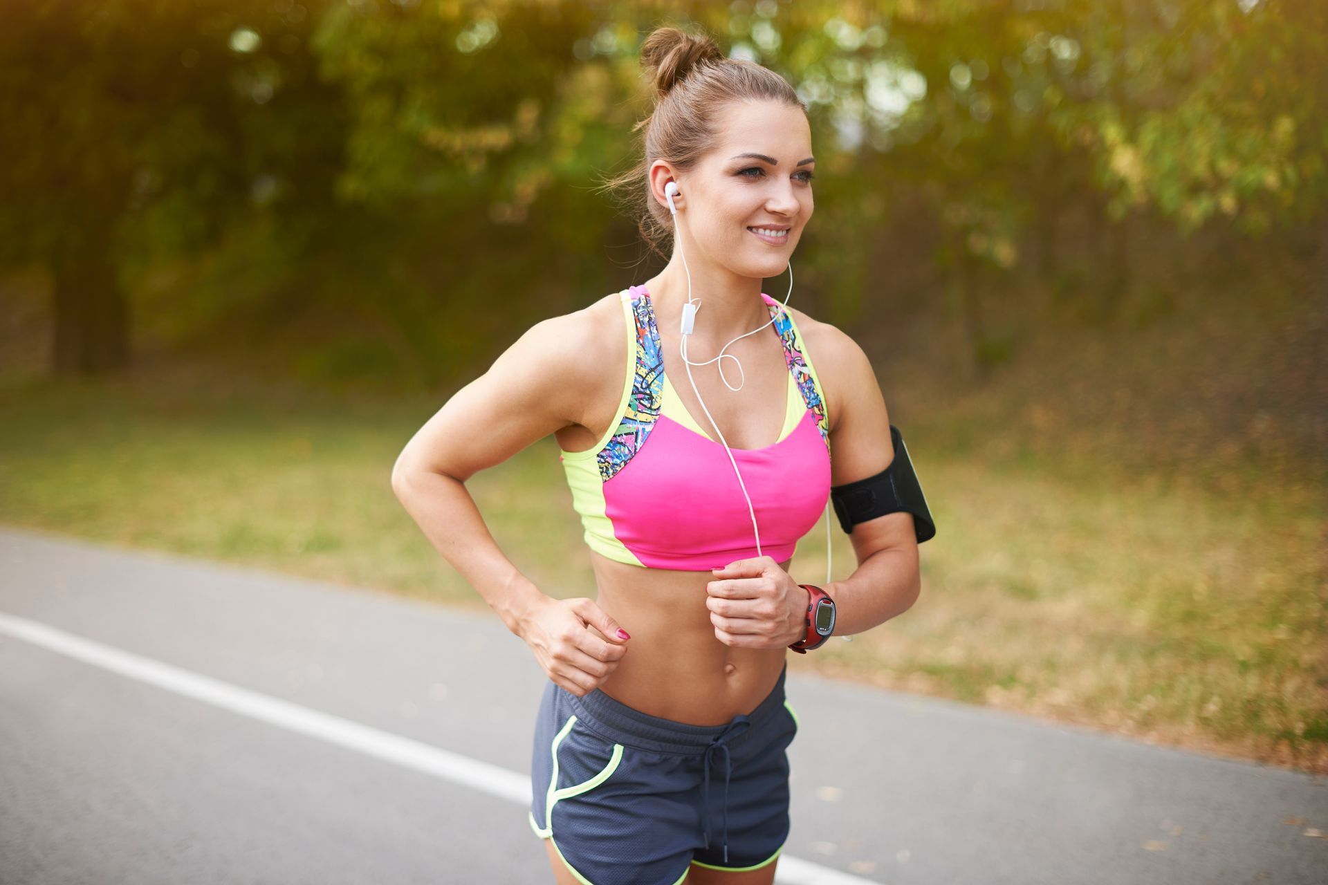 Woman running on a path, wearing pink and blue athletic wear, smiling, with earbuds and a phone armband.