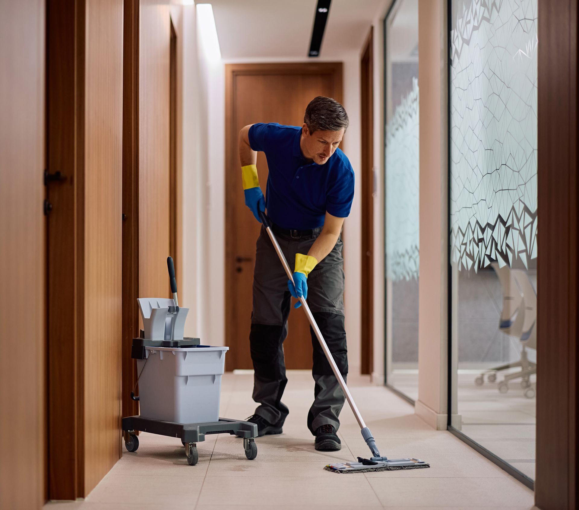 Janitor mopping hallway floor beside cleaning cart and supplies in a modern office building