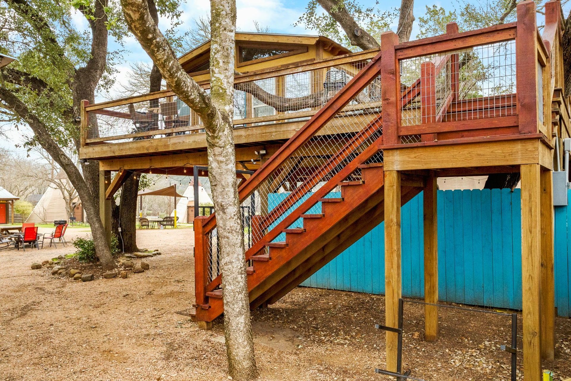 Wooden treehouse with stairs and a balcony, set in a yard with trees and blue fence.