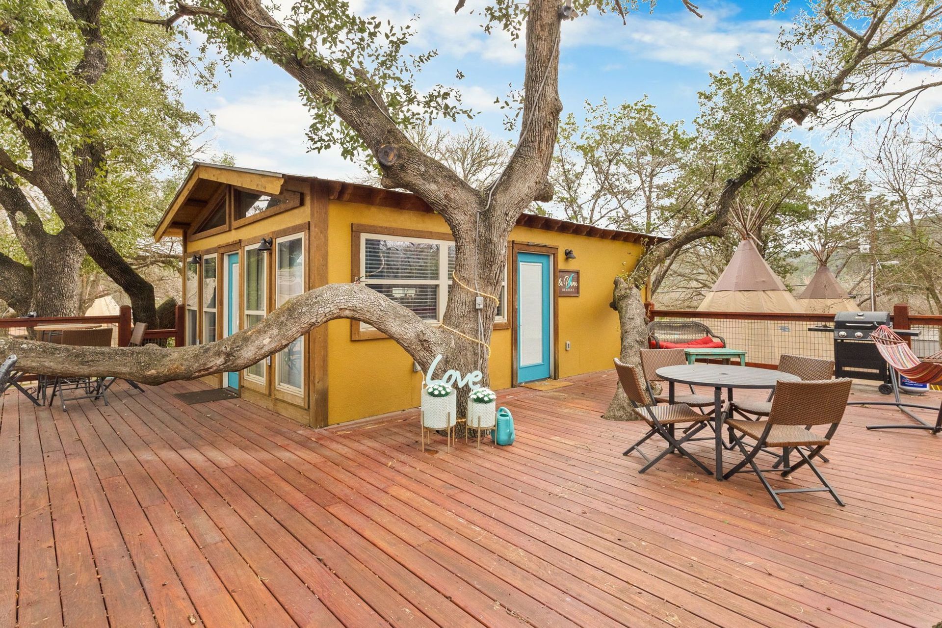 A yellow cabin with a turquoise door on a wooden deck, tree branches overhead, outdoor seating.