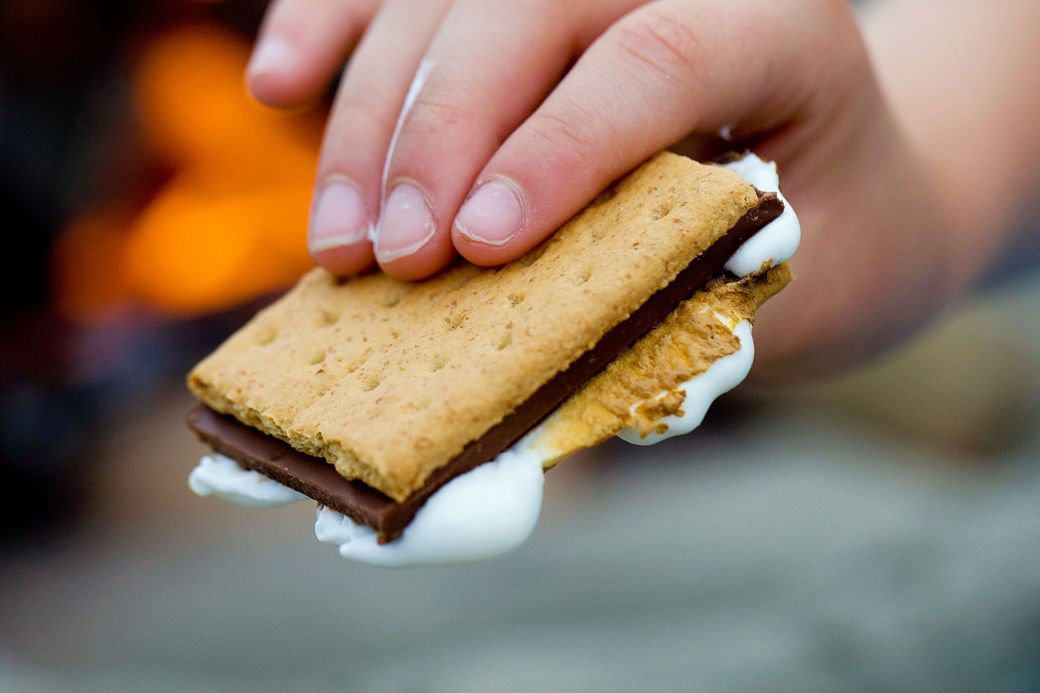 Hand holding a s'more, with melted marshmallow and chocolate between graham crackers; campfire in background.