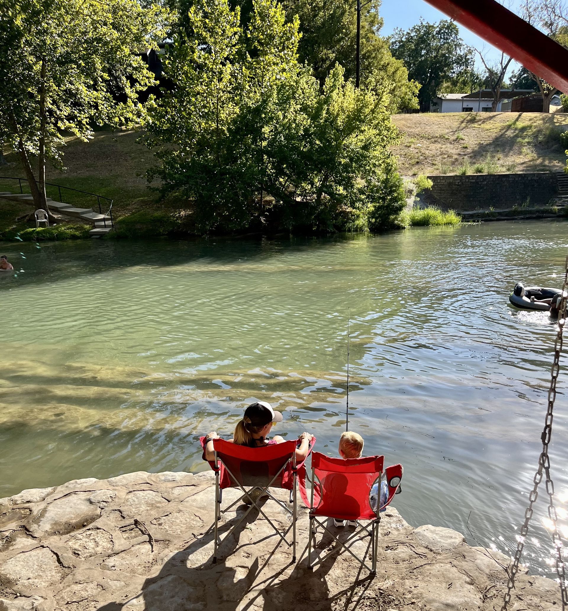 Two people sit in red chairs, near a clear river. Sunny day.