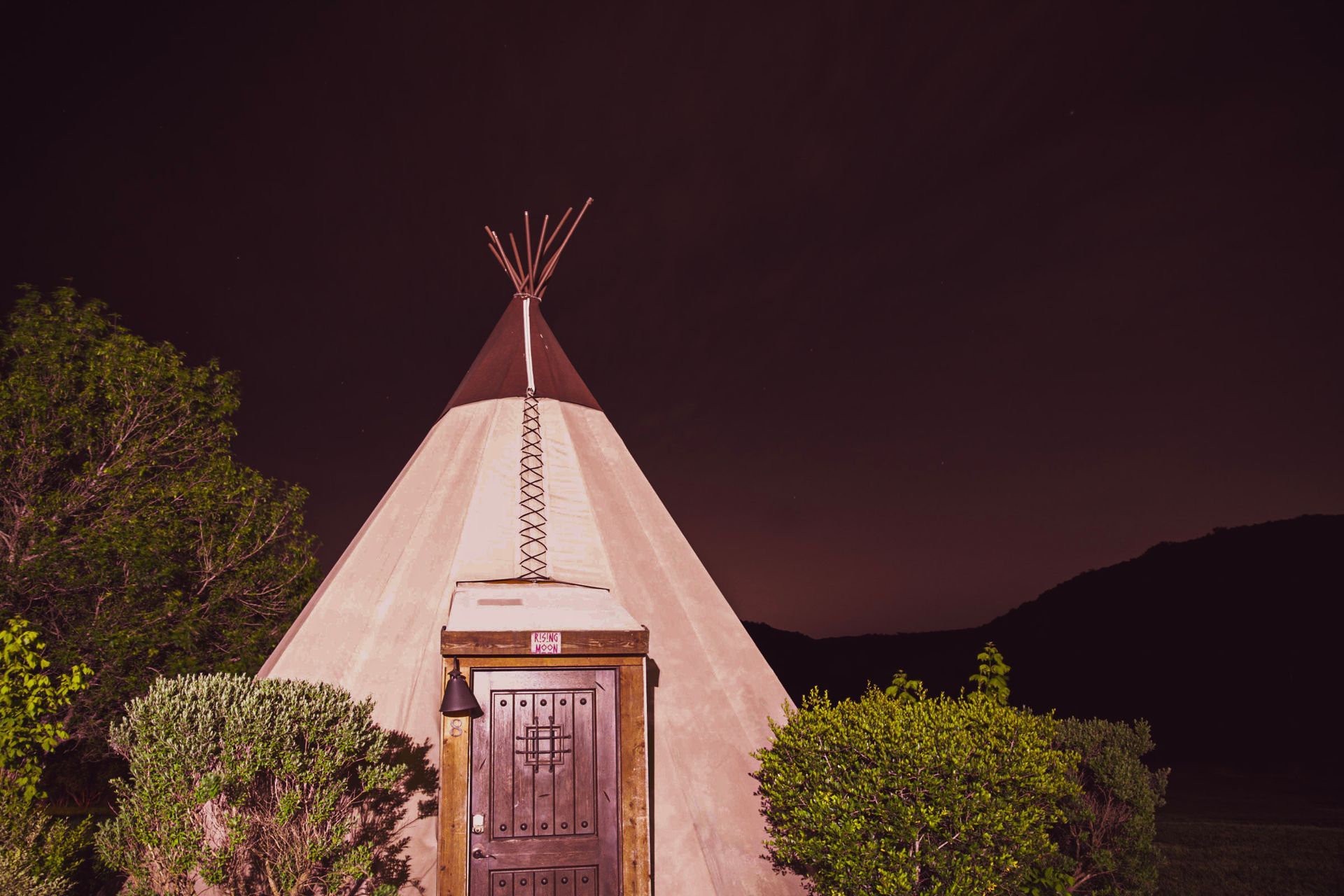 A tipi with a wooden door, illuminated at night. Bushes frame the entrance, mountains in the distance.