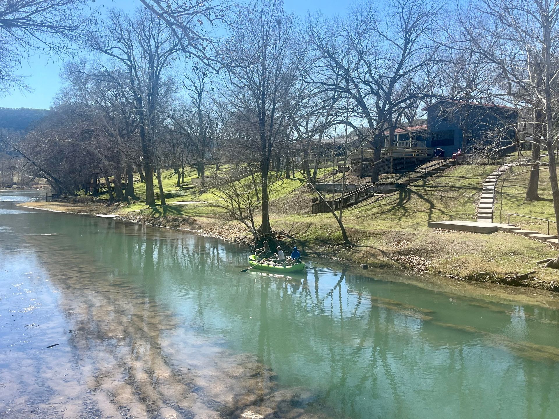 River with kayakers near a grassy bank and trees on a sunny day.