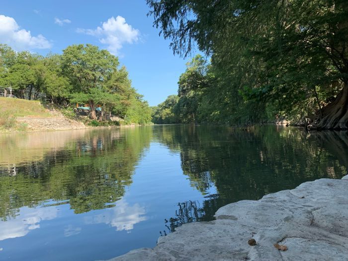 Calm river flowing between trees with sky reflection; rock in foreground.
