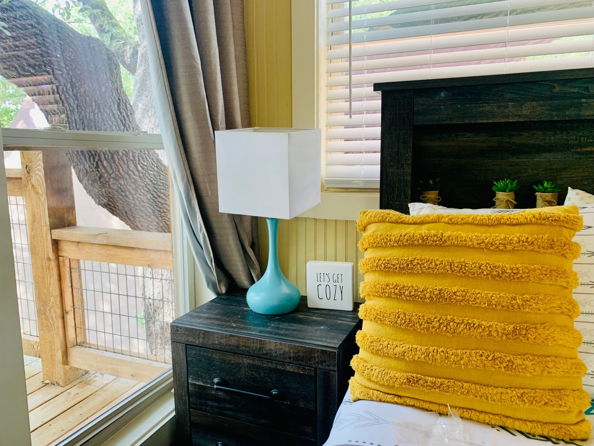 Bedroom with teal lamp, yellow pillow, and a window overlooking a wooden deck and tree.