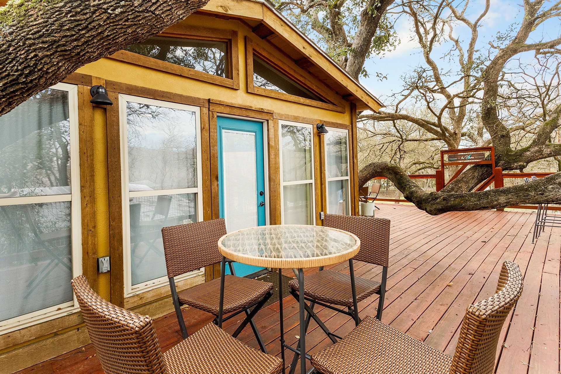 Wooden deck with table and chairs outside a small cabin with a blue door, nestled among tree branches.