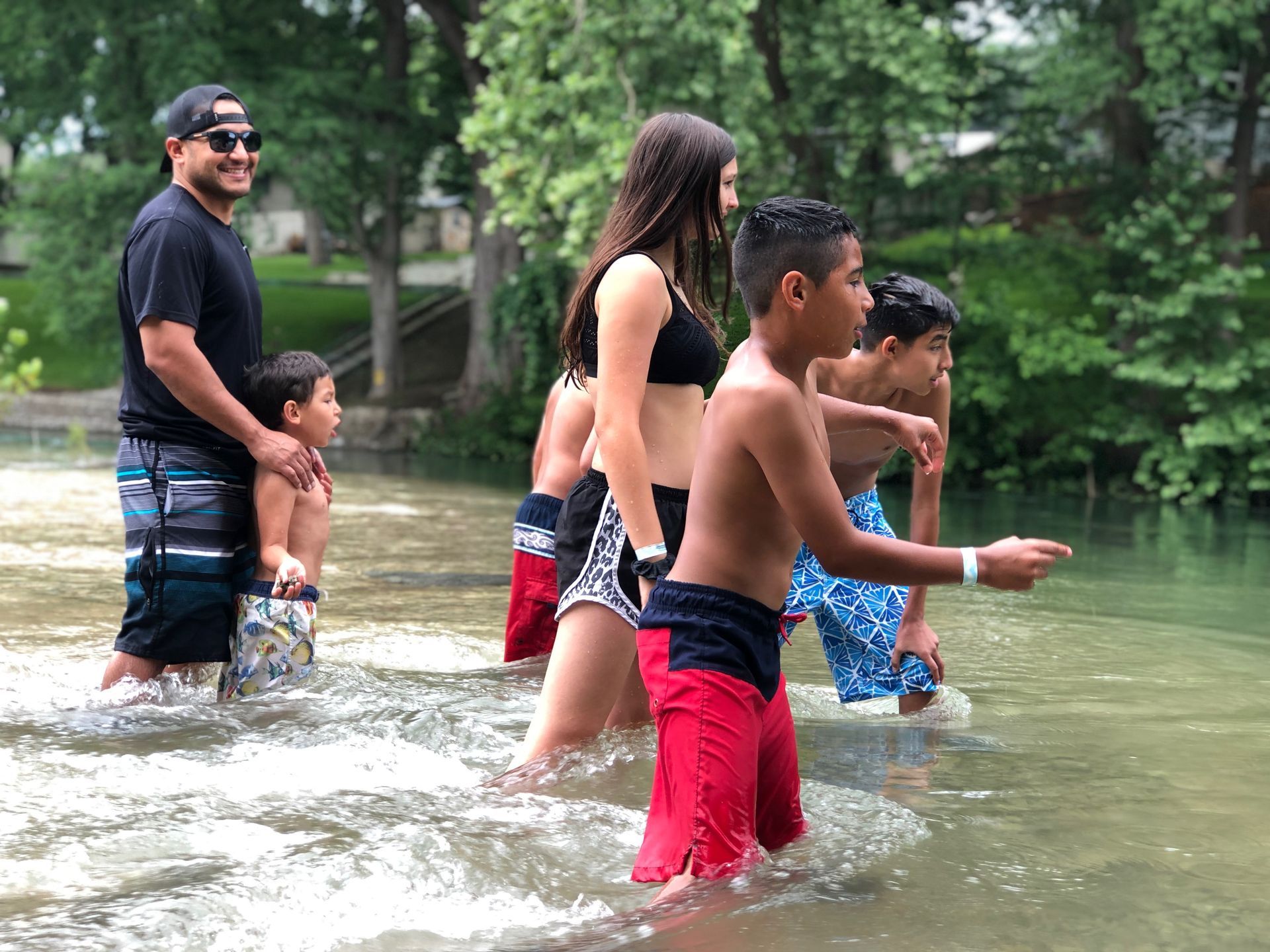 Family wading in a river, splashing water. Person in sunglasses holds a child. Other kids in swimwear. Trees in background.