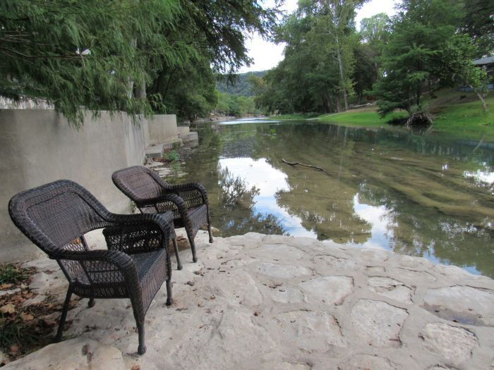 Two wicker chairs sit on a rocky riverbank, overlooking a calm, tree-lined river.