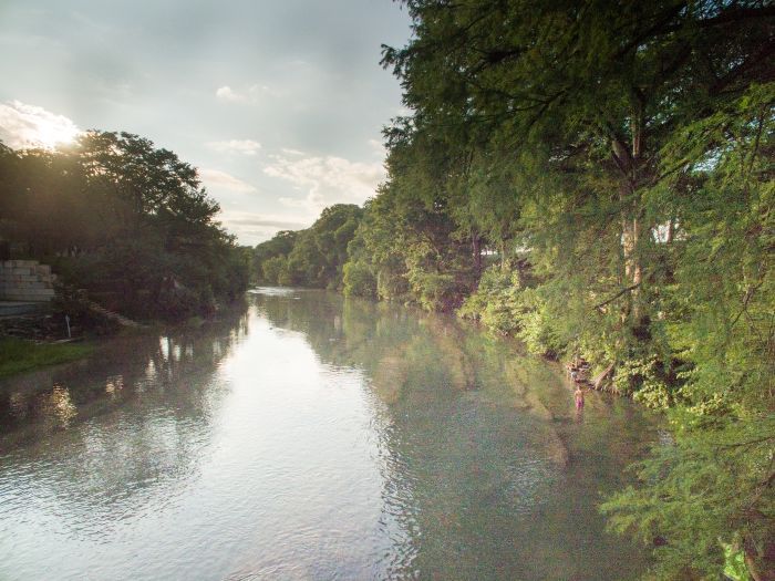 River flowing through a tree-lined bank under a cloudy sky; sunlight peeks through the trees.