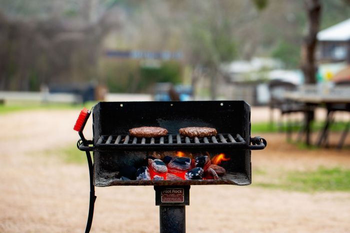 Burgers grilling on a charcoal grill outdoors with trees and picnic tables blurred in the background.