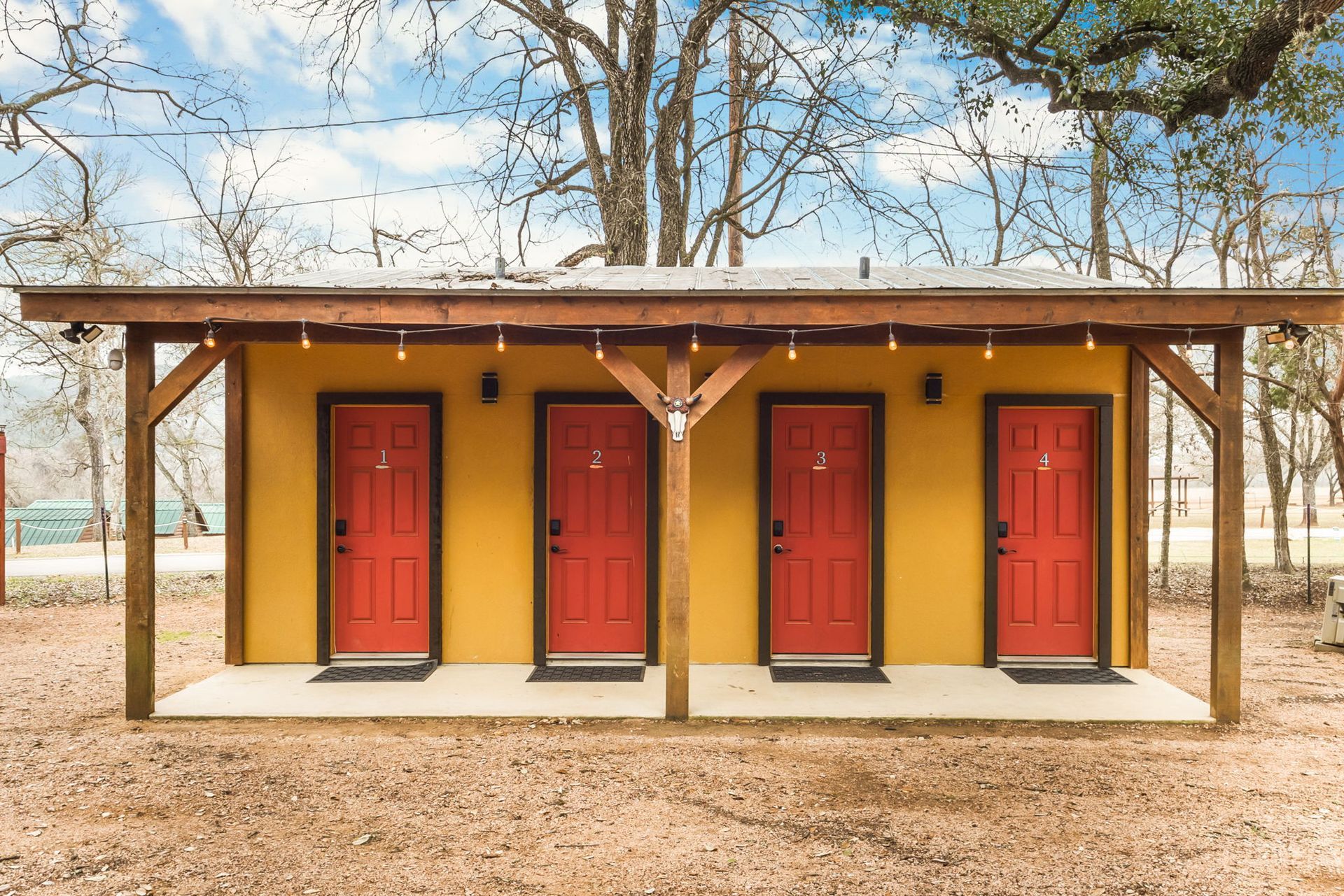 Yellow building with four red doors under a brown wooden awning, set in a wooded area.