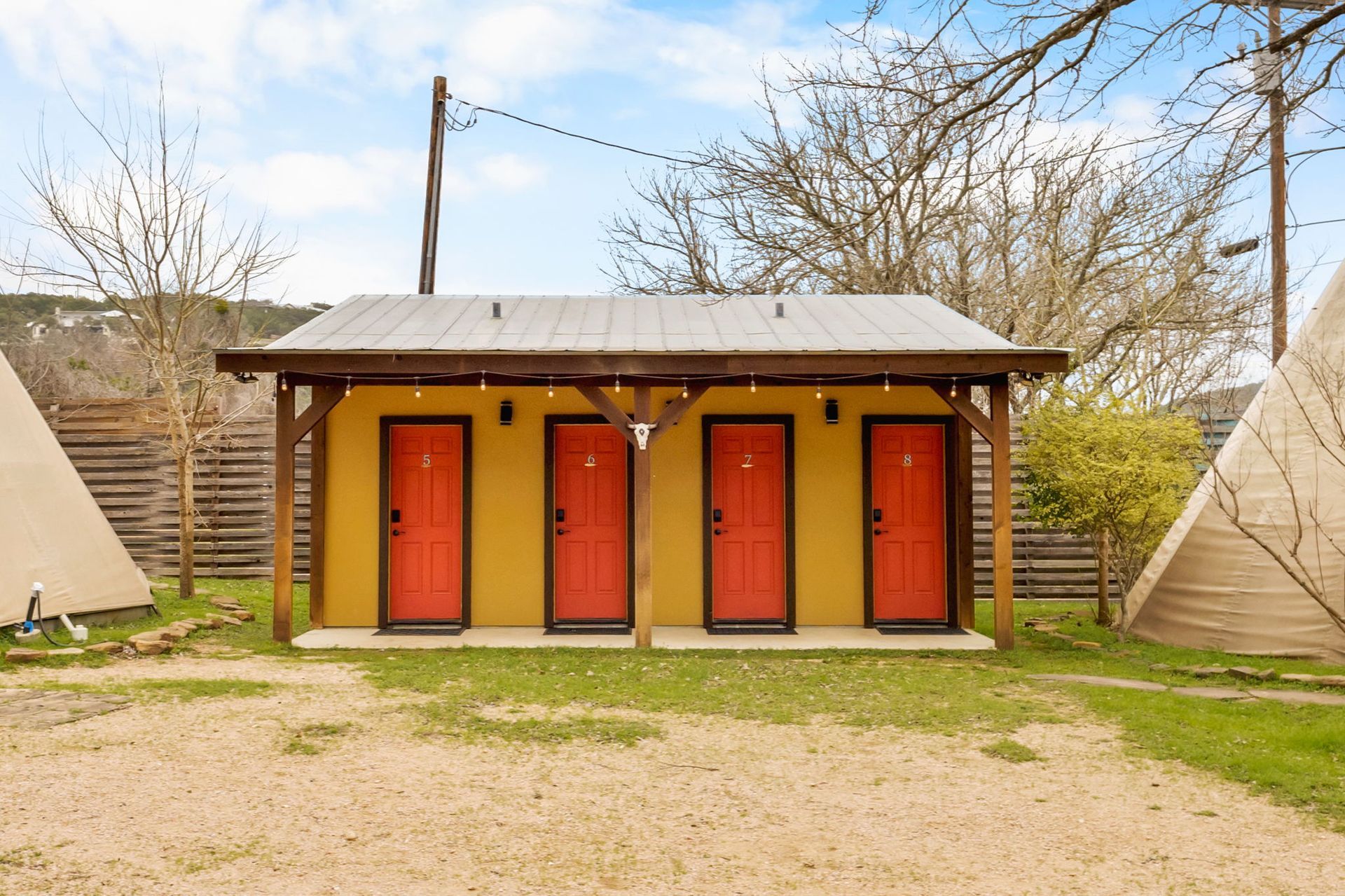 Yellow building with four red doors, under a brown roof, flanked by teepees, set in a grassy outdoor area.