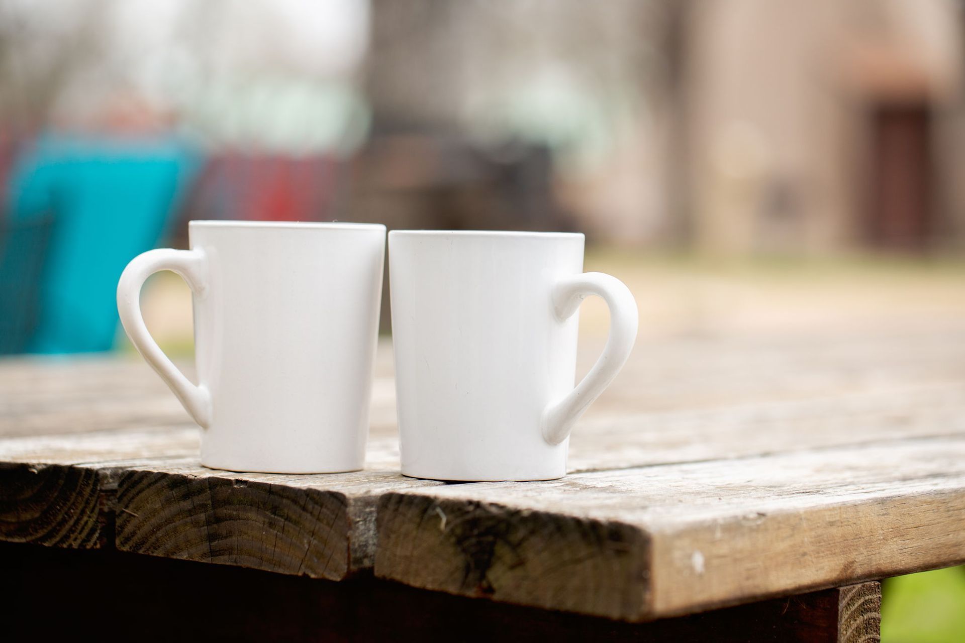 Two white coffee mugs on a weathered wooden table, set outdoors with blurred background.