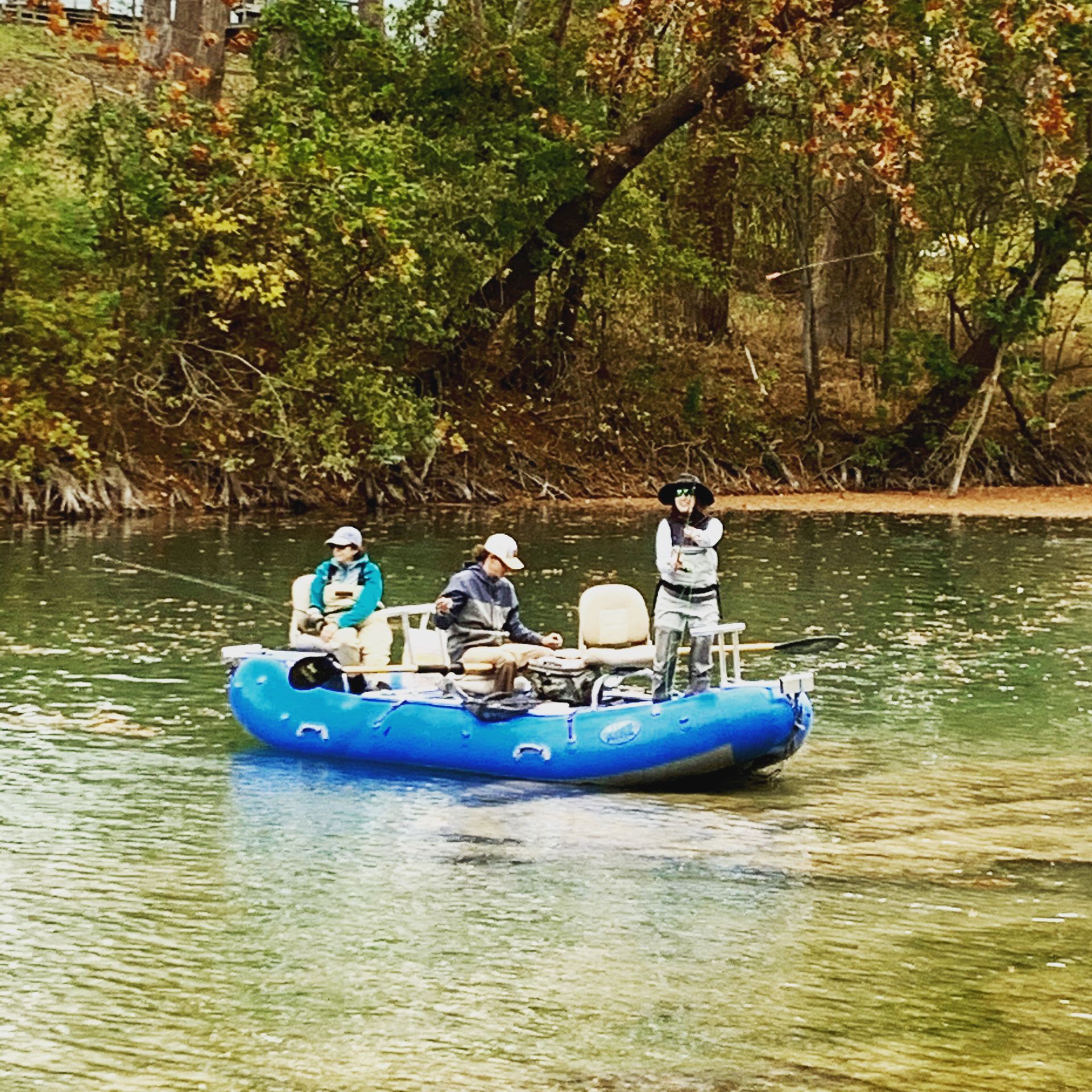 Three people fly fishing from a blue raft on a river with green trees.