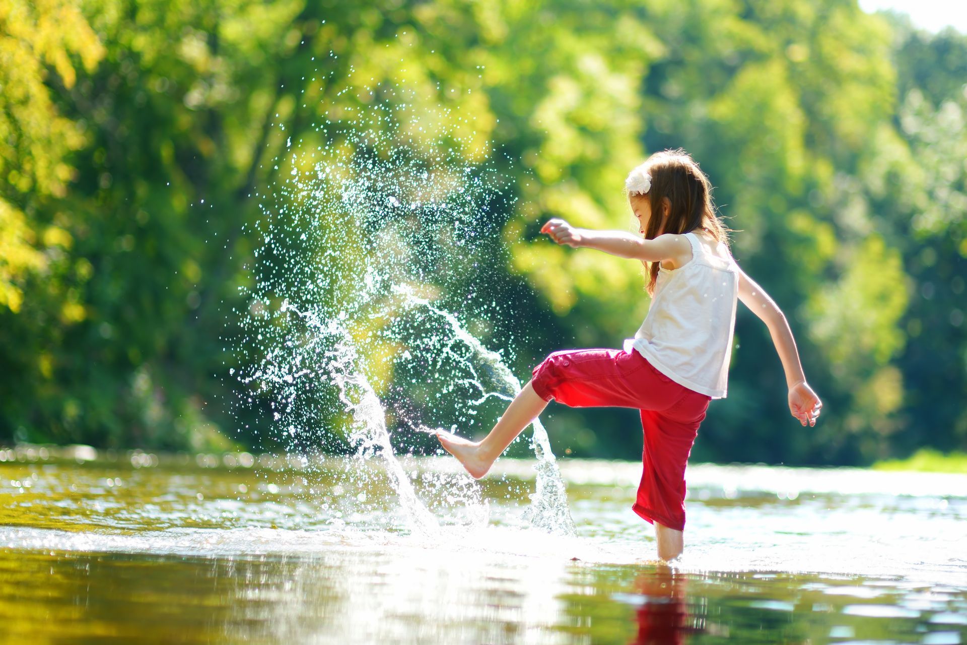Girl splashing in a shallow river, red pants, white shirt, sunny day, green trees in background.