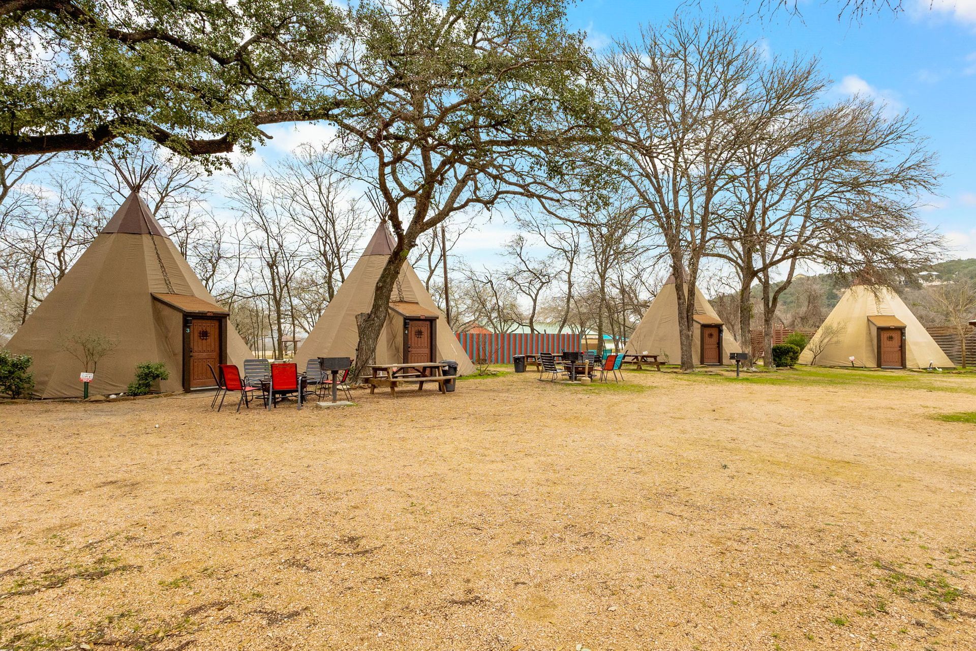 Teepees in a row with picnic tables and chairs on a gravel lot under trees.