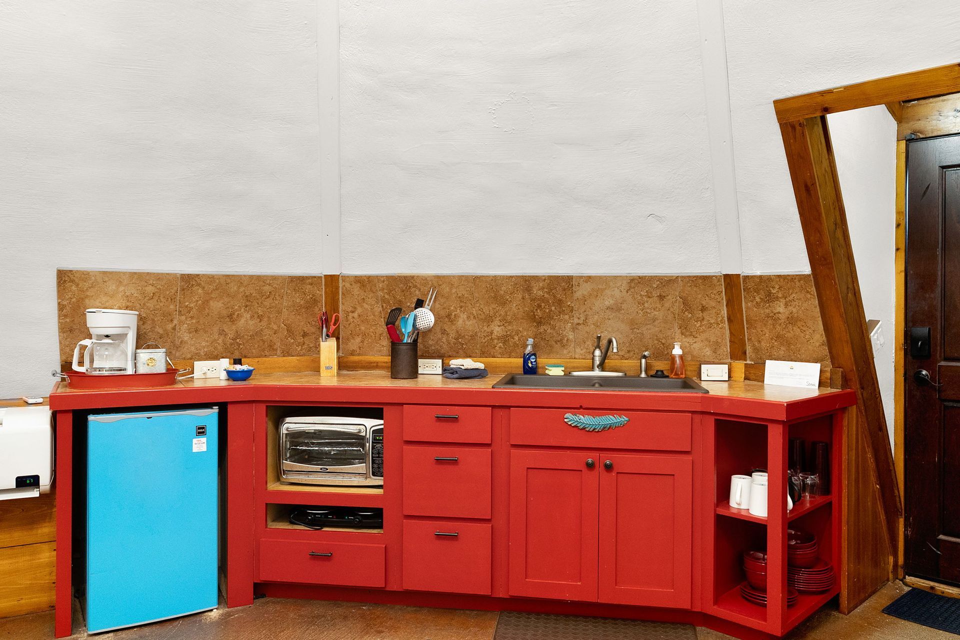 Red kitchen cabinets with sink, microwave, mini-fridge, and door.