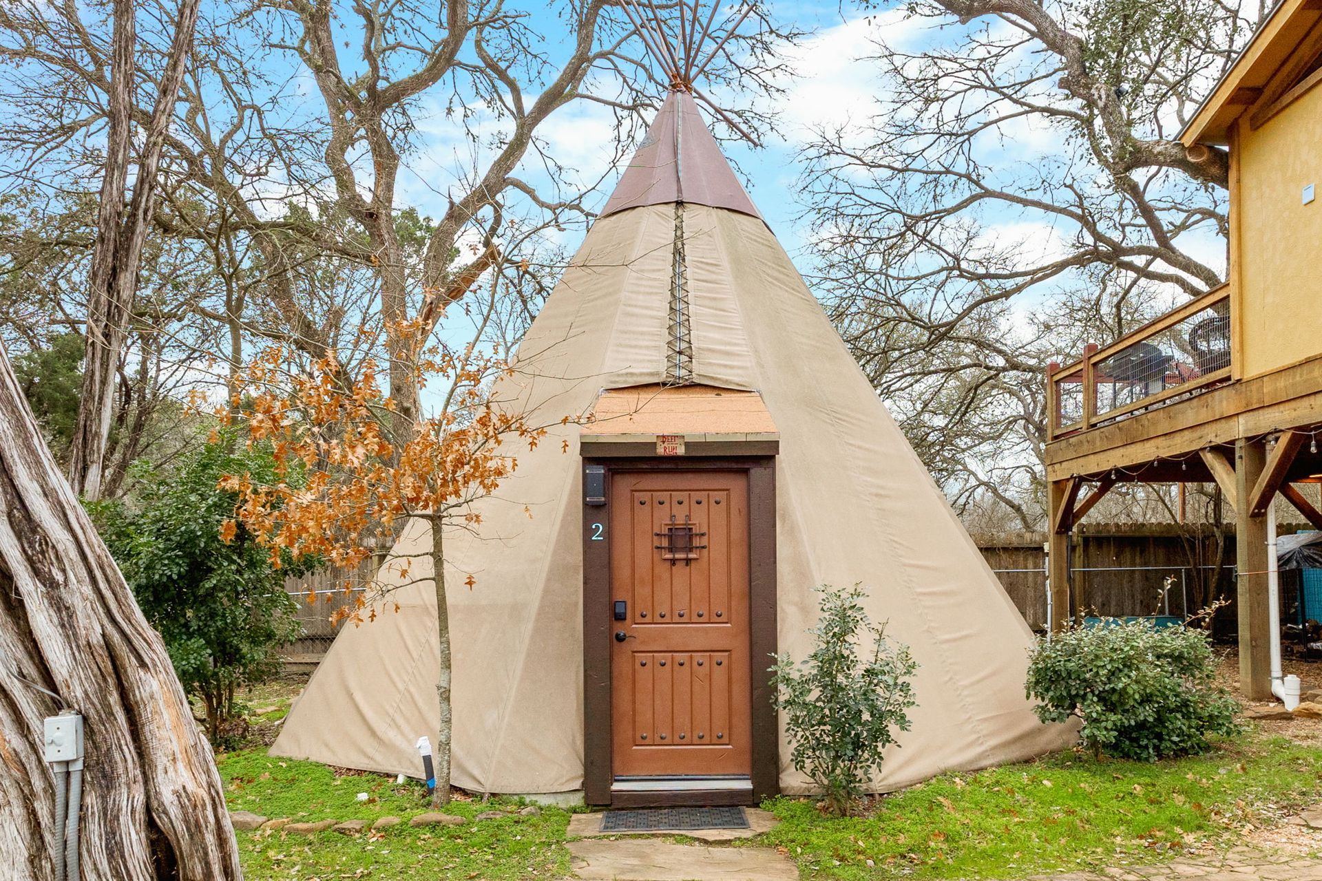 Tan tipi with brown door and a small porch, set in a grassy yard, with a wooden deck to the right.