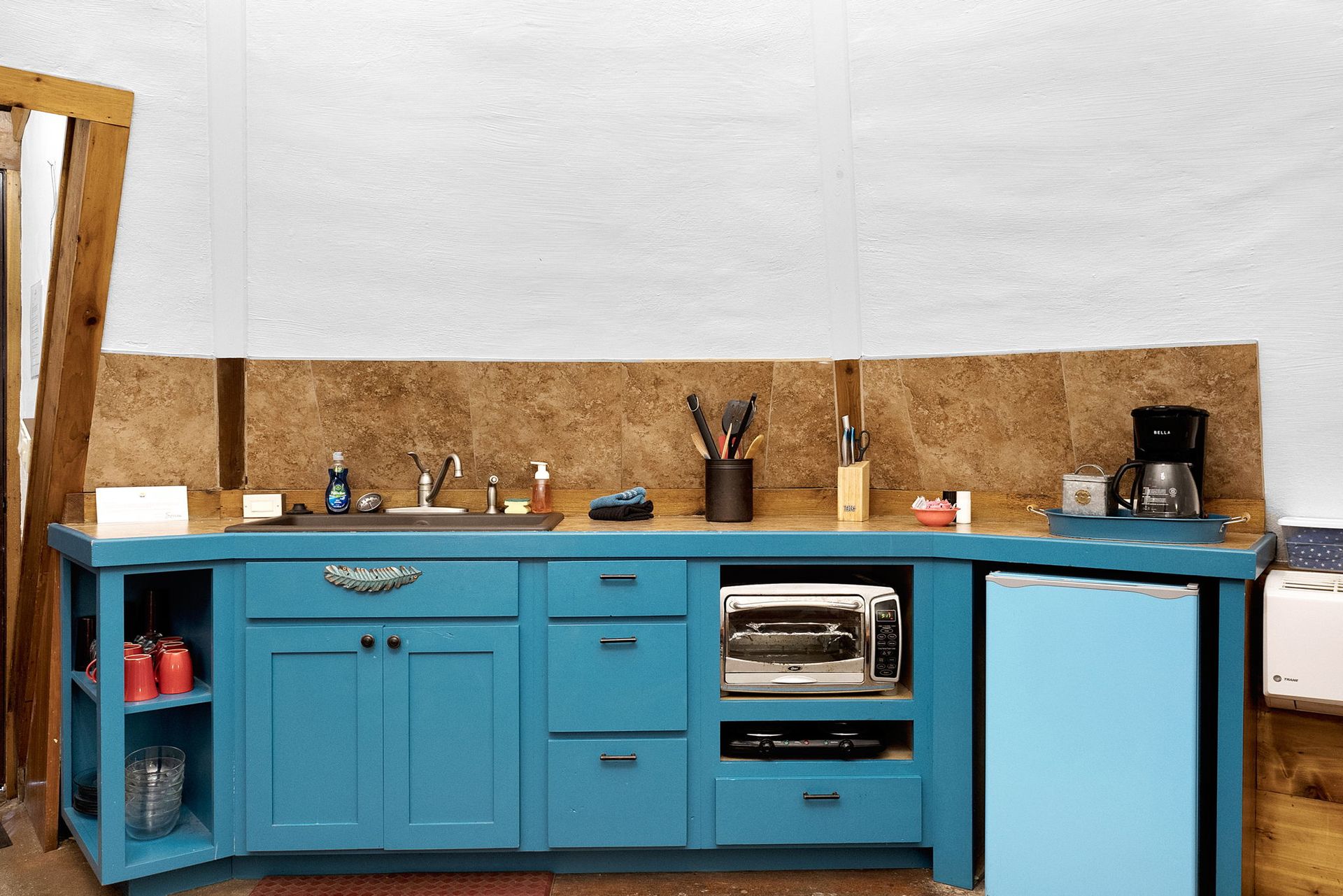 Blue kitchen cabinets with sink, microwave, and small refrigerator. Brown countertop and cork backsplash.