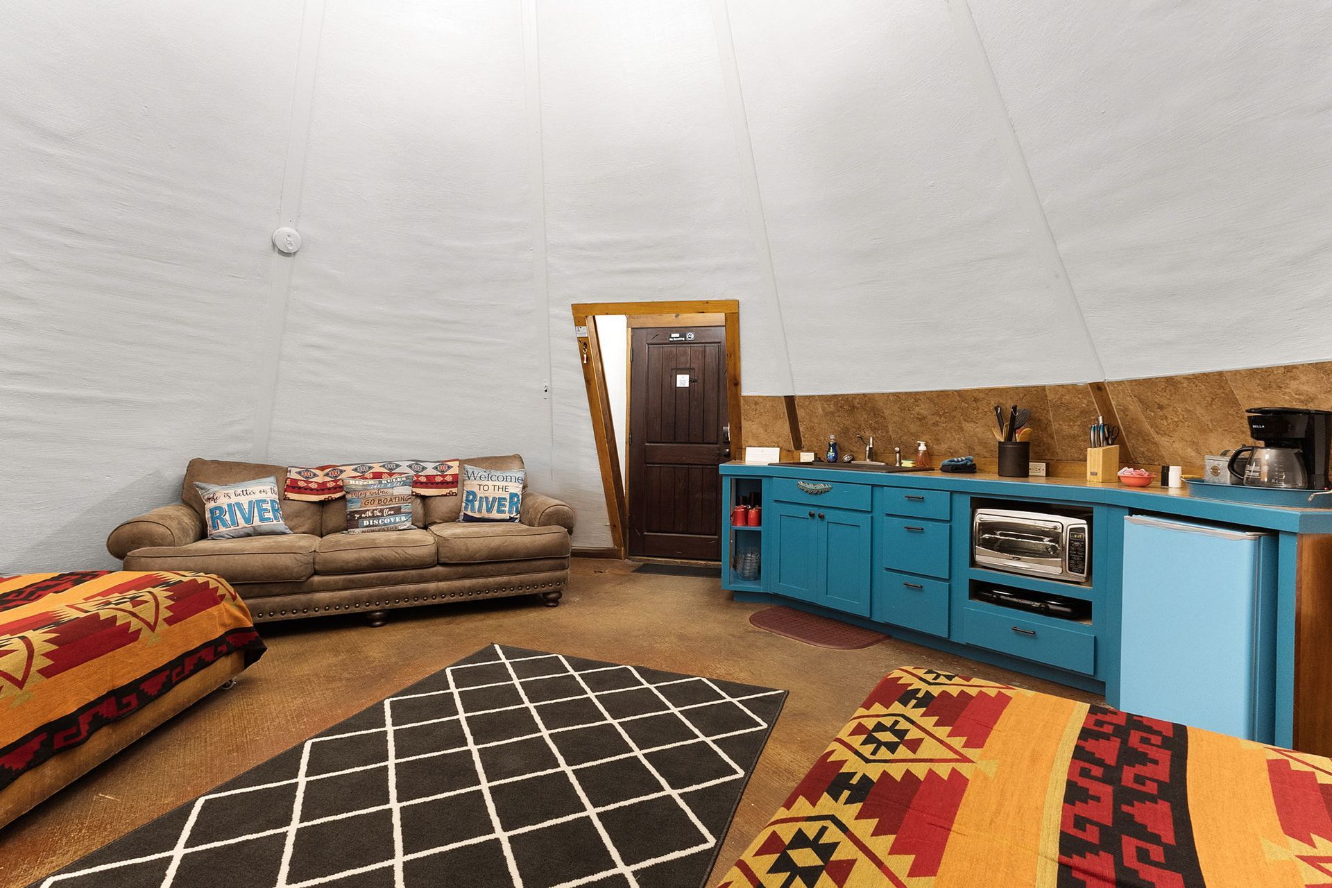 Interior of a dome-shaped room with a kitchen, sofa, and floor cushions. Bright blue cabinetry, light walls.
