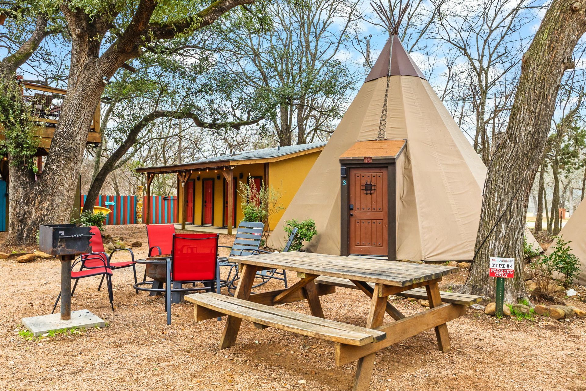 Campground scene with picnic table, grill, and teepee next to a building under trees.