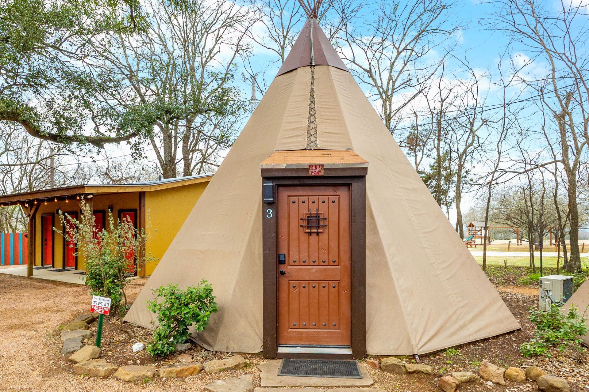 Tan teepee-shaped structure with brown door, set outdoors with other buildings and trees.