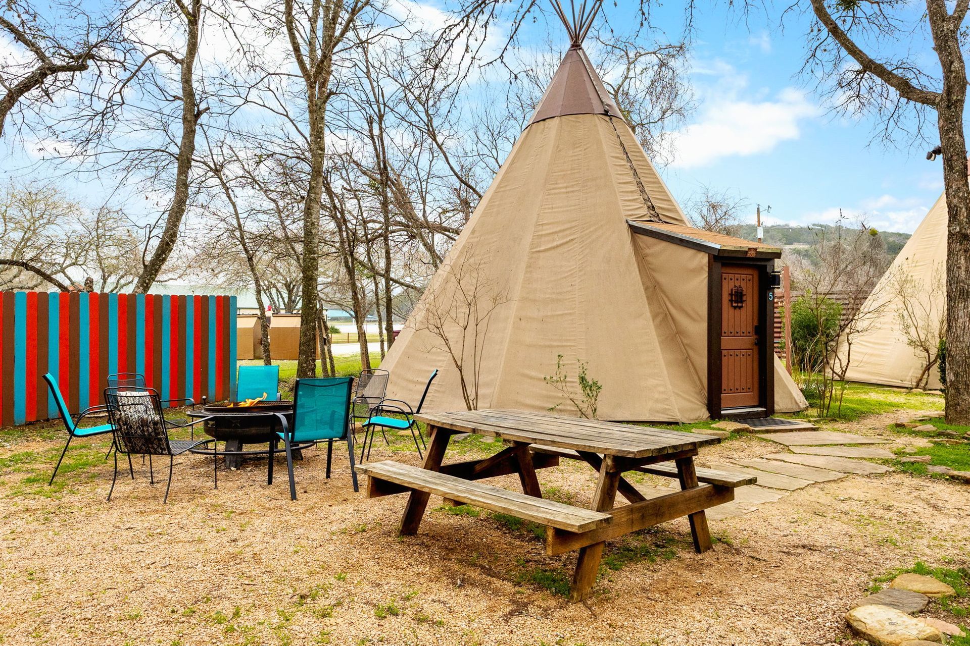 A tan teepee with a picnic table, fire pit, and blue chairs in a yard.