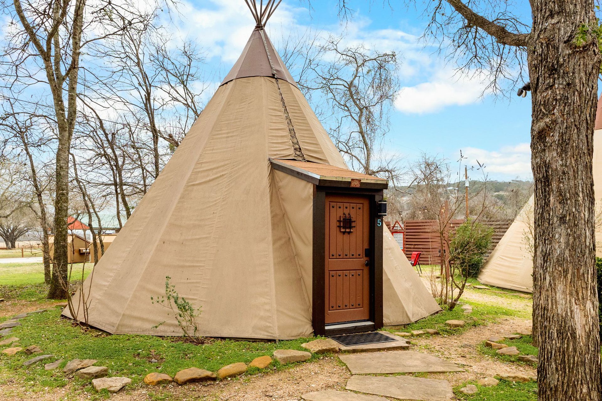 Tan teepee with a brown door, on a grassy plot. Stone path and trees in the background.