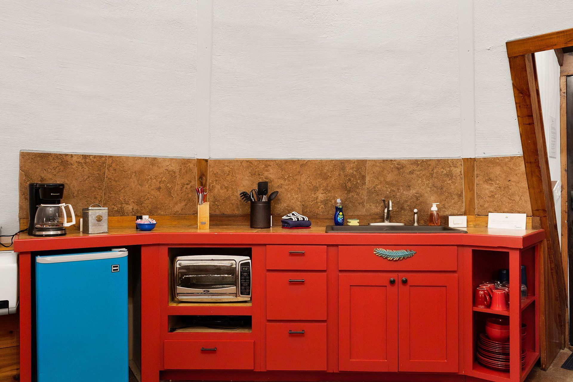 Red kitchen with blue mini-fridge, toaster oven, sink, and various items on the countertop.