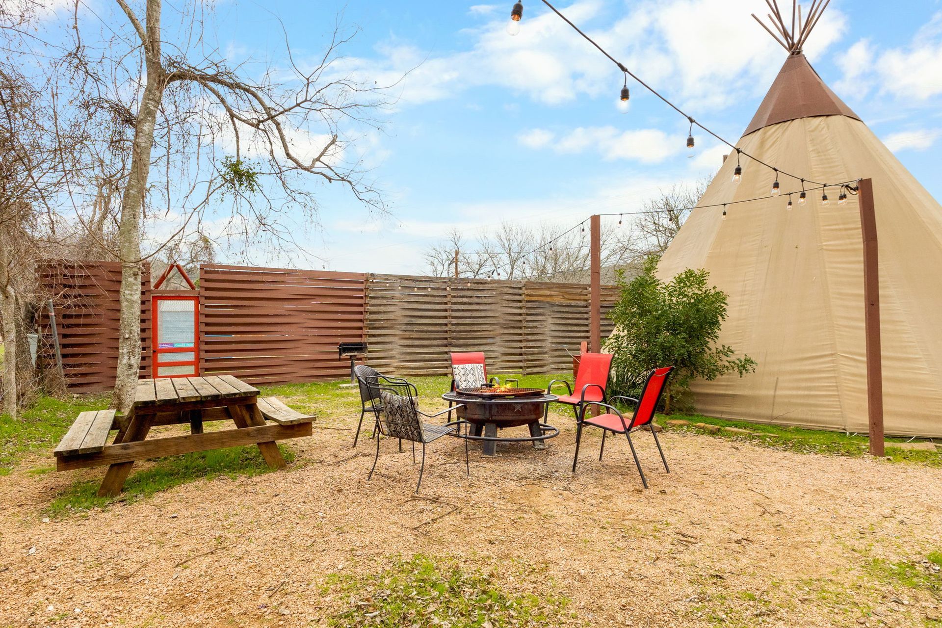 Backyard with a teepee, picnic table, fire pit, and string lights, set on gravel with a wooden fence.